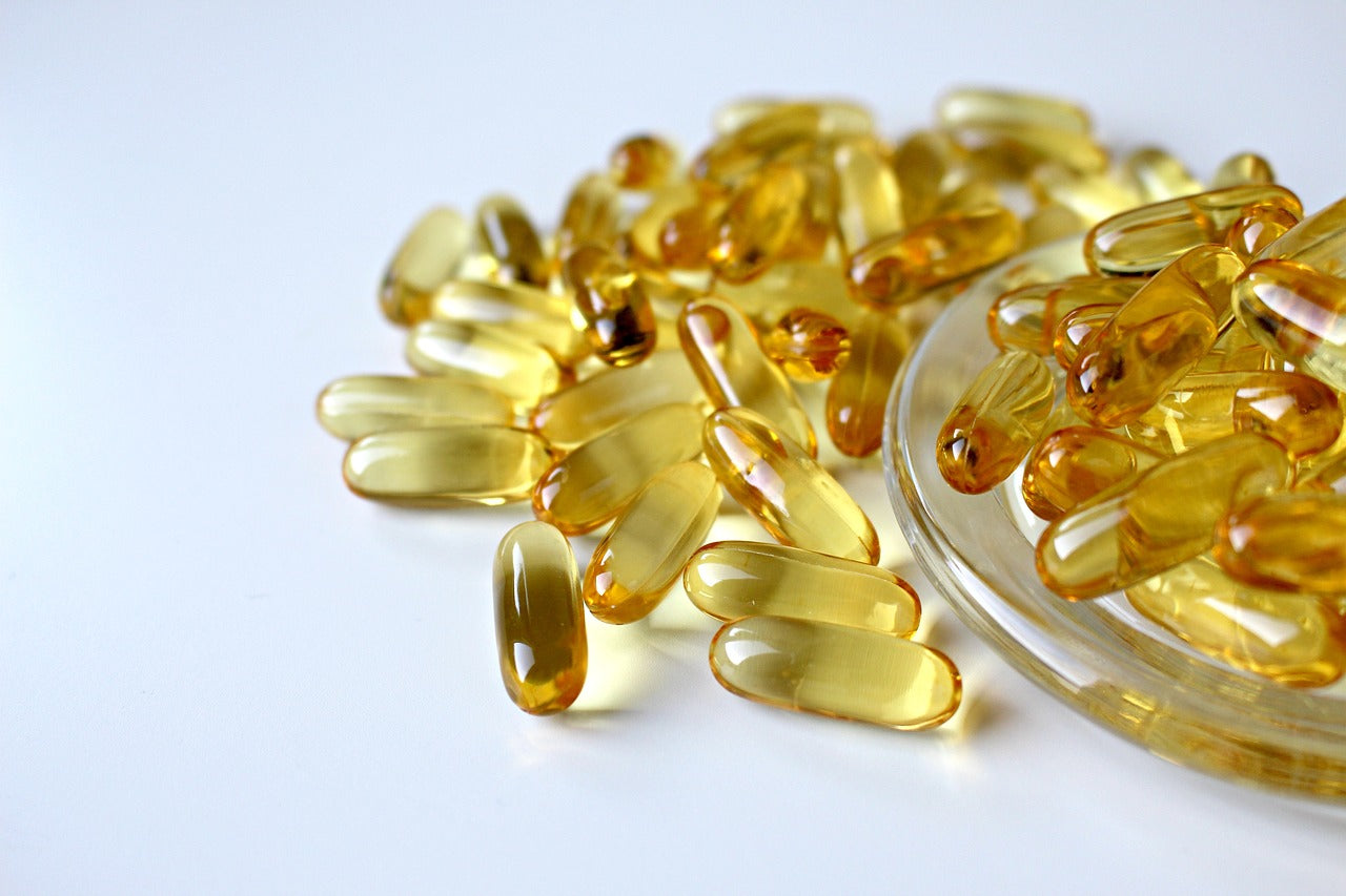 Capsules dorées d’oméga 3 dispersées sur une surface blanche, avec une coupelle en verre remplie de compléments alimentaires.
