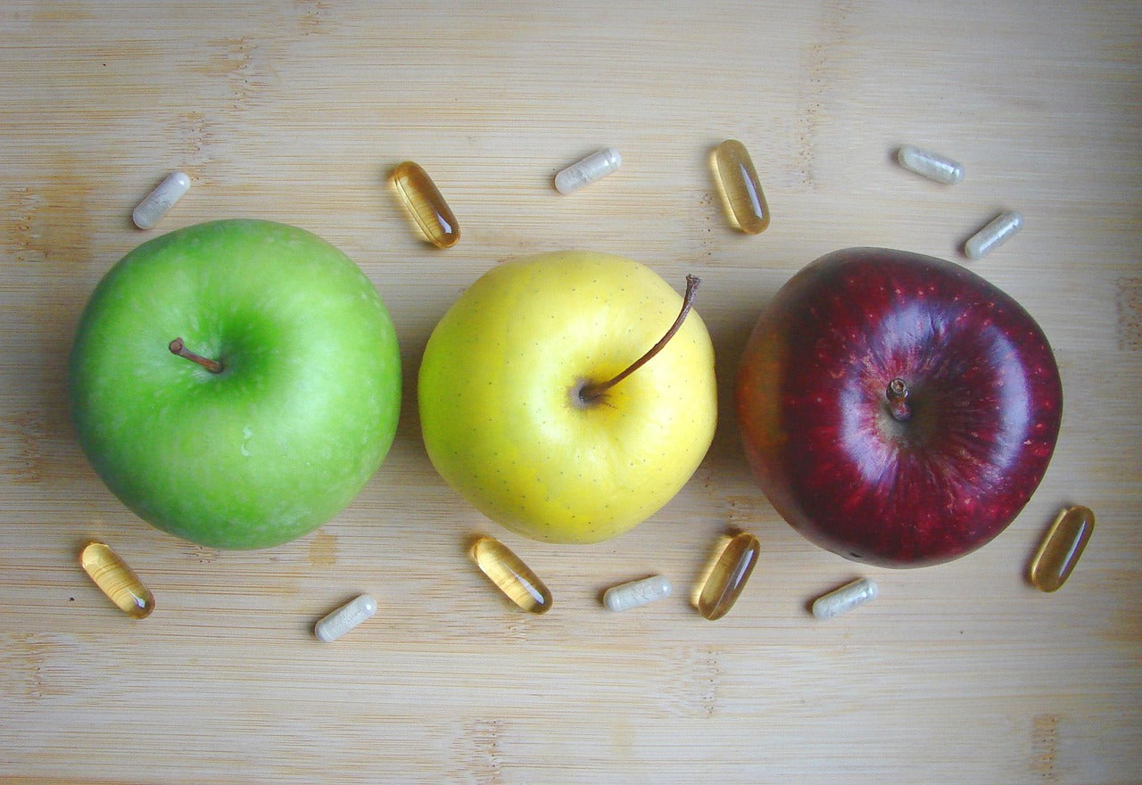 Trois pommes alignées sur une planche en bois, entourées de capsules et gélules de compléments naturels.