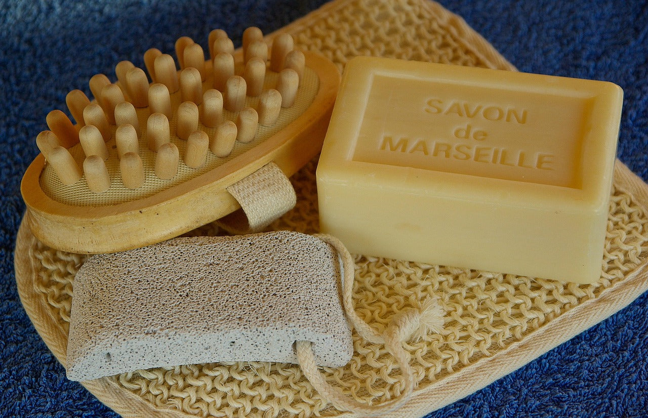 Accessoires de soin naturels comprenant un savon de Marseille, une brosse en bois et une pierre ponce posés sur une serviette.
