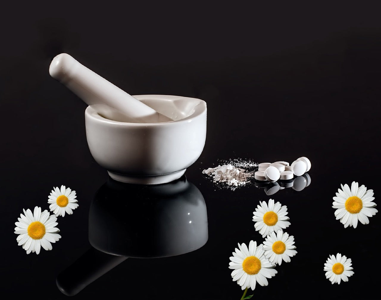 Mortier et pilon blancs avec des comprimés écrasés et des fleurs de camomille, symbolisant la préparation d’élixirs naturels pour l’équilibre hormonal.