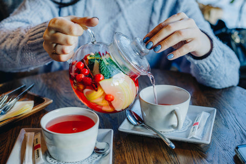 Théière en verre remplie d’infusion aux fruits et plantes, versée dans une tasse pour soutenir les besoins féminins comme le SPM ou la digestion.